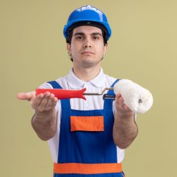 young builder man in construction uniform and safety helmet demonstrating paint roller looking at camera with serious face standing over green background young builder man in construction uniform and safety helmet demonstrating paint roller looking at camera with serious face standing over green background